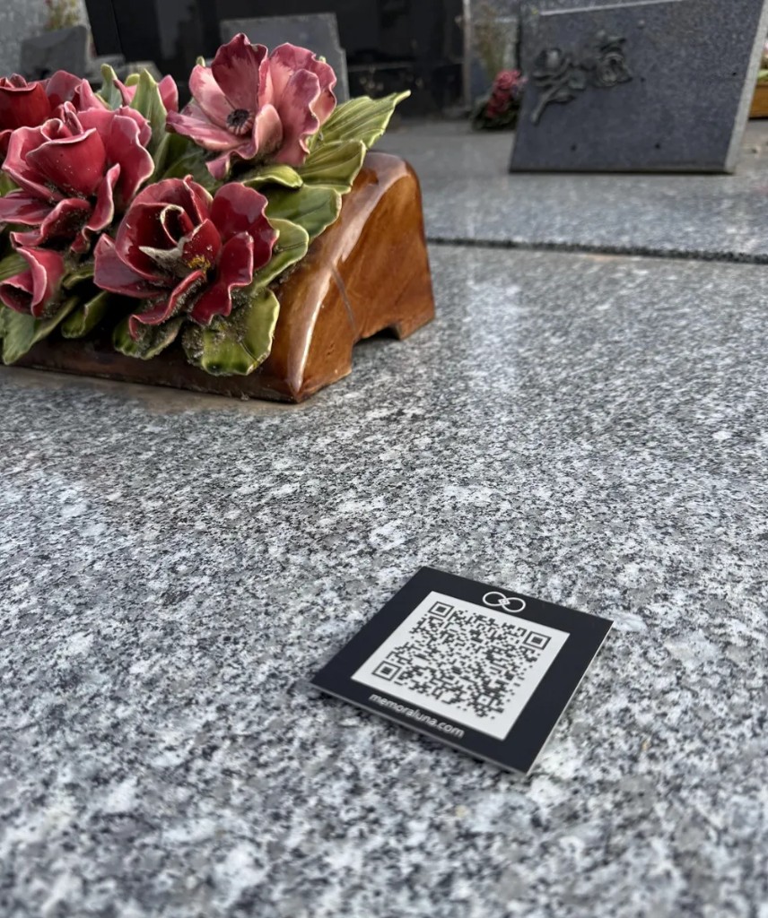 Plaque QR code Memoraluna posée sur le granit d'une sépulture, à côté d'un bouquet de fleurs.